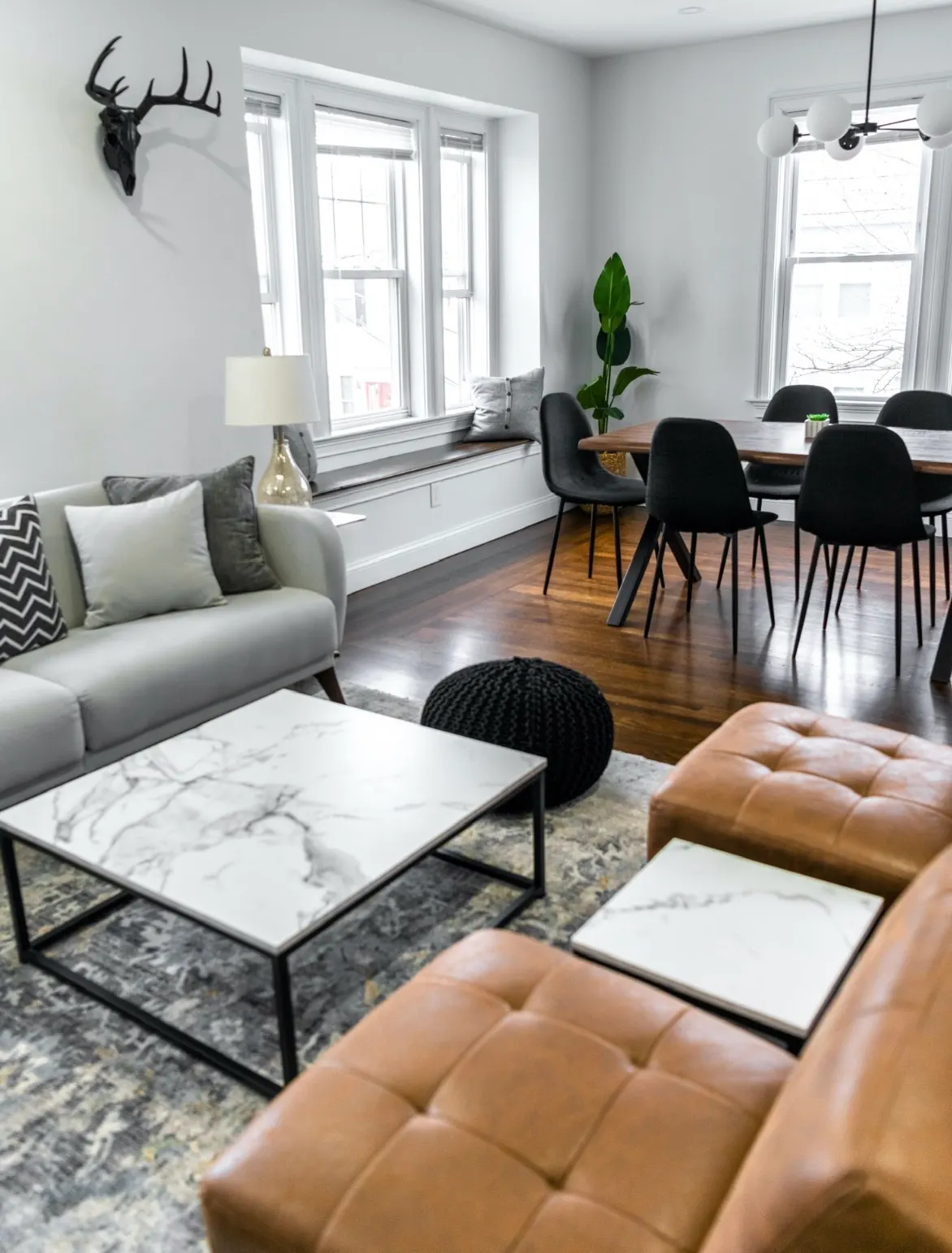 Modern living room and dining area with light grey walls, dark wood floors, marble coffee table, and tan ottomans.