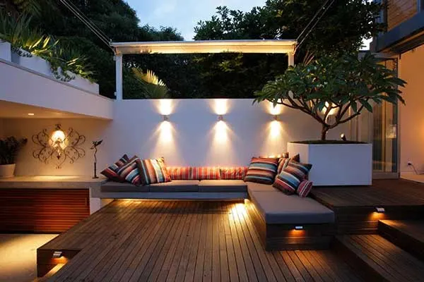 Modern outdoor patio with wooden decking, built-in sectional seating, decorative multicolored pillows, and ambient lighting.