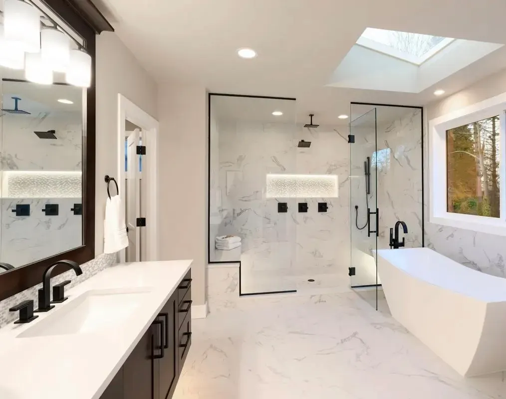 Modern bathroom with white marble tile, large walk-in shower, freestanding tub, dark vanity, black fixtures, and skylight.