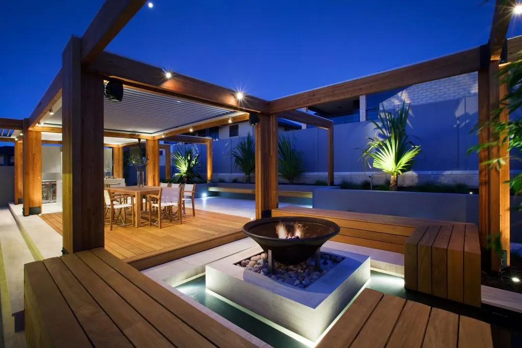 Luxurious outdoor living area features a modern pergola, fire pit, outdoor kitchen, and ambient lighting at dusk.