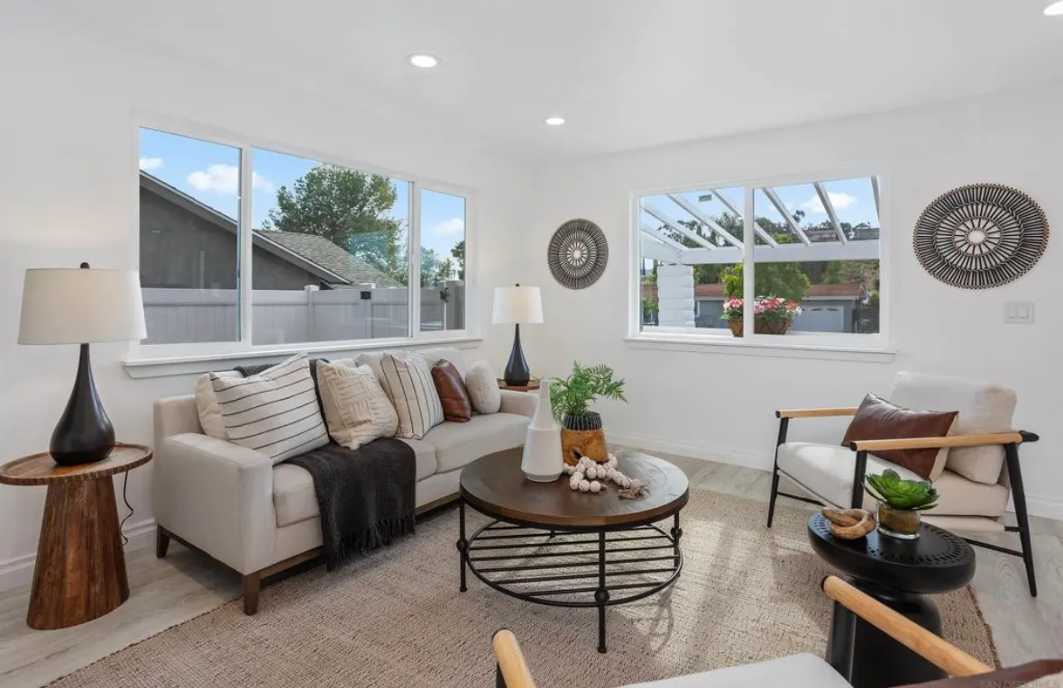 Bright living room with white walls, cozy neutral furniture, large windows, and a round wooden coffee table.