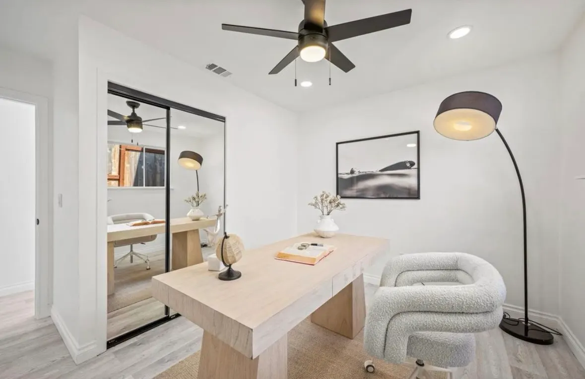 Modern home office with light wood desk, soft boucle chair, black ceiling fan, and mirrored closet doors.