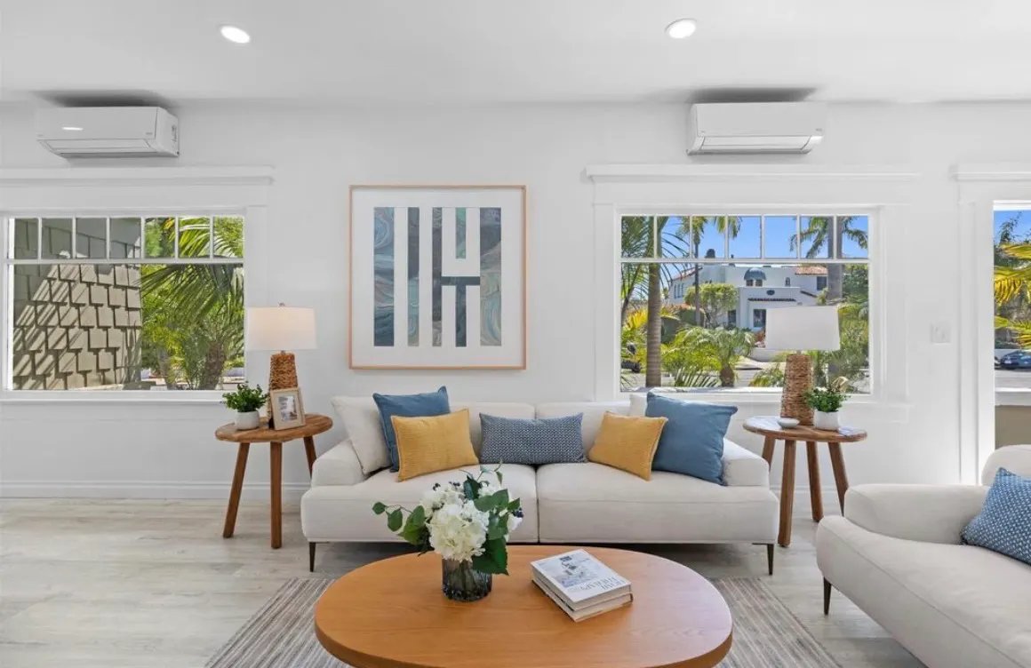 Bright, modern living room with a white sofa, yellow and blue pillows, light wood flooring, and large windows.