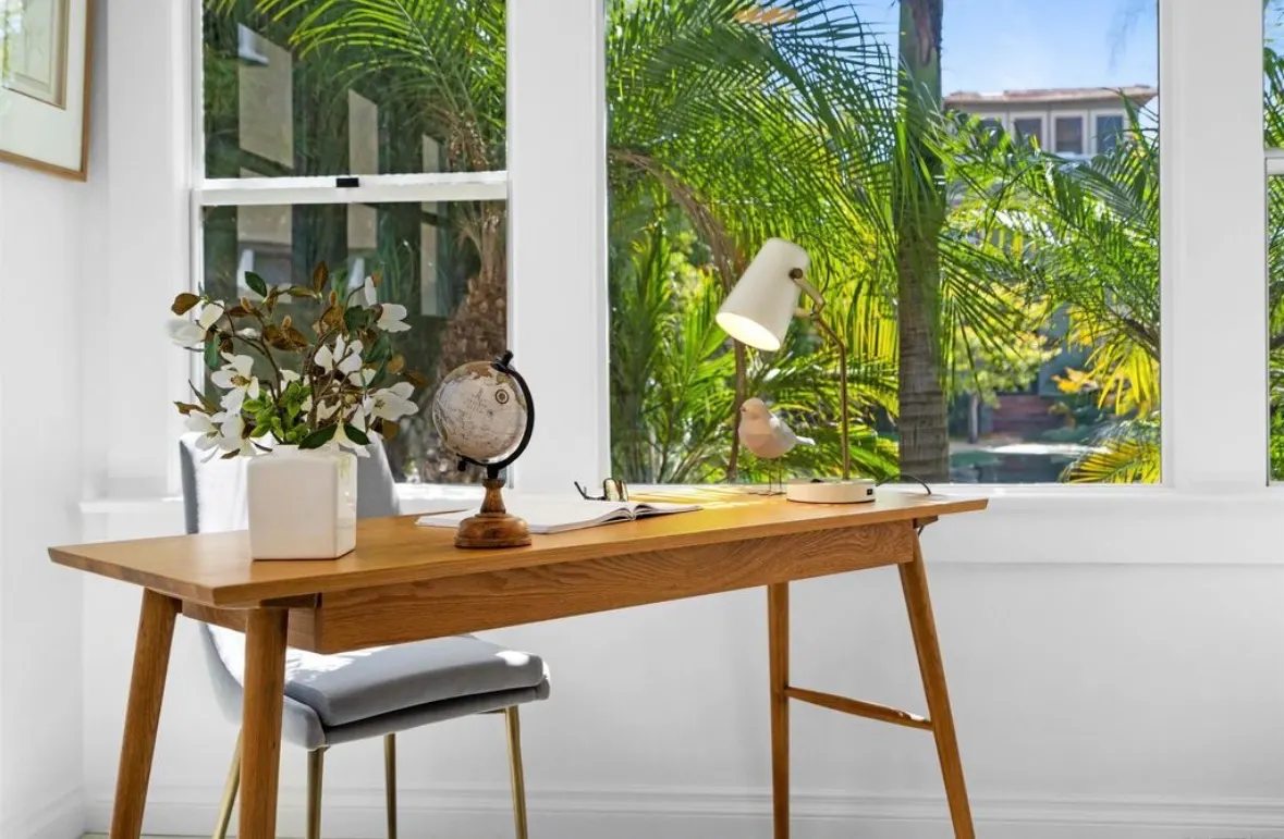 Bright home office with natural light, a wood desk, white wall, gray chair, and tropical views.