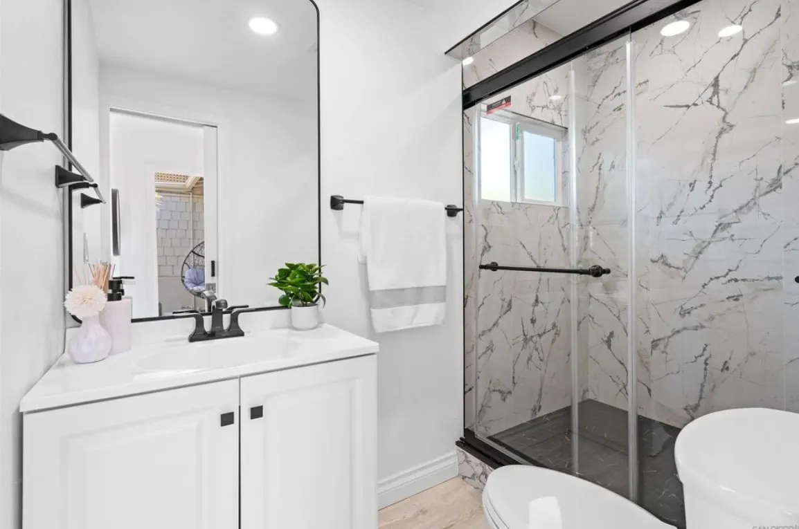 Modern bathroom with white vanity, black framed mirror, marble-patterned shower, and sleek black fixtures.