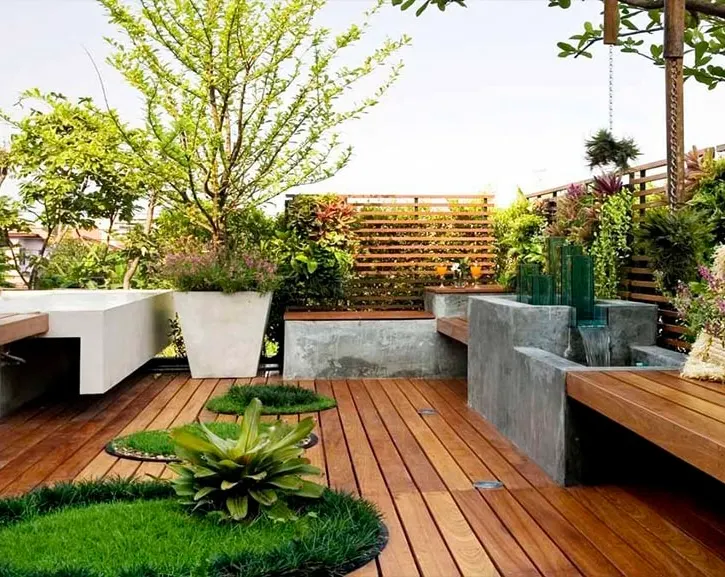Modern rooftop deck with rich wooden flooring, built-in concrete benches, lush plants, and a water feature.