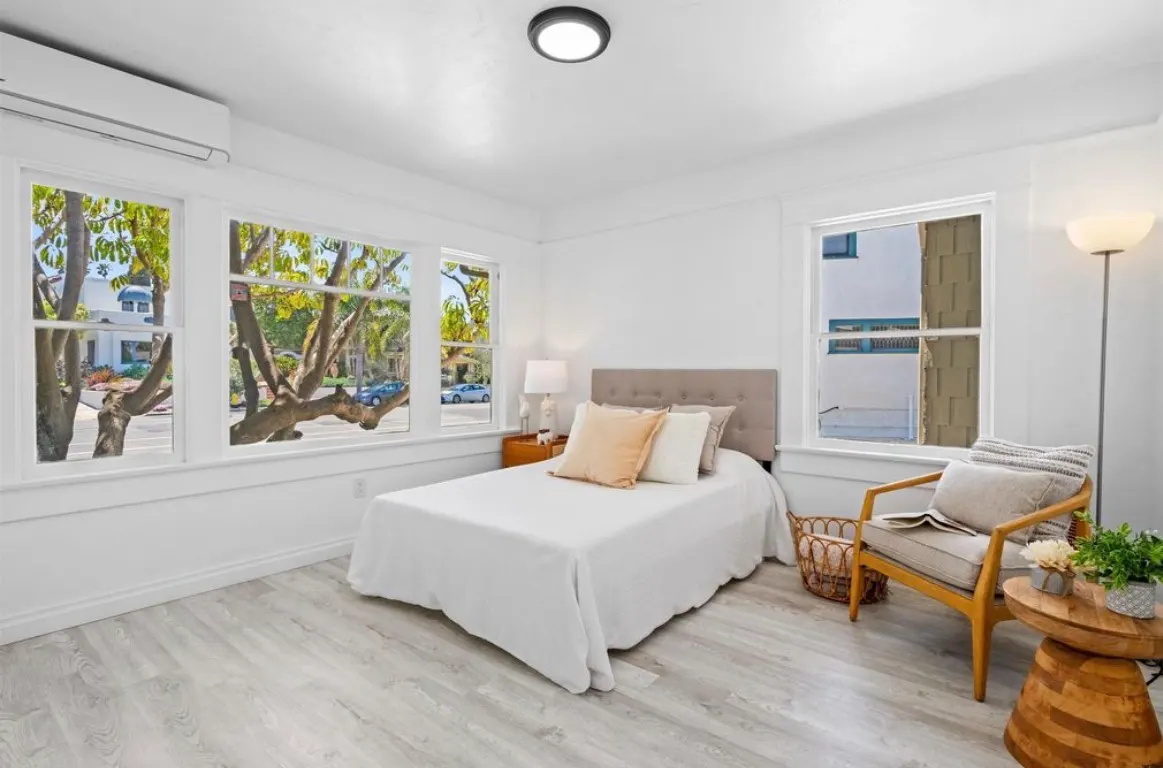 Brightly remodeled bedroom with white walls, light wood flooring, large windows, and simple modern furnishings.