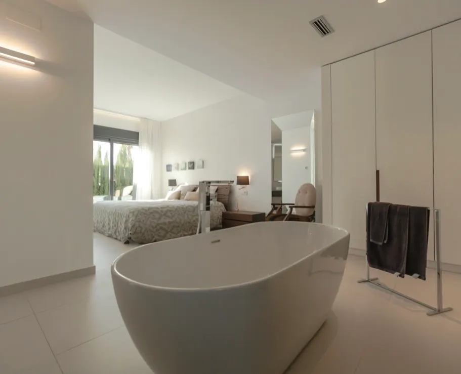 Modern open-plan master bathroom and bedroom with crisp white tones, freestanding tub, and seamless entry to an outdoor patio.