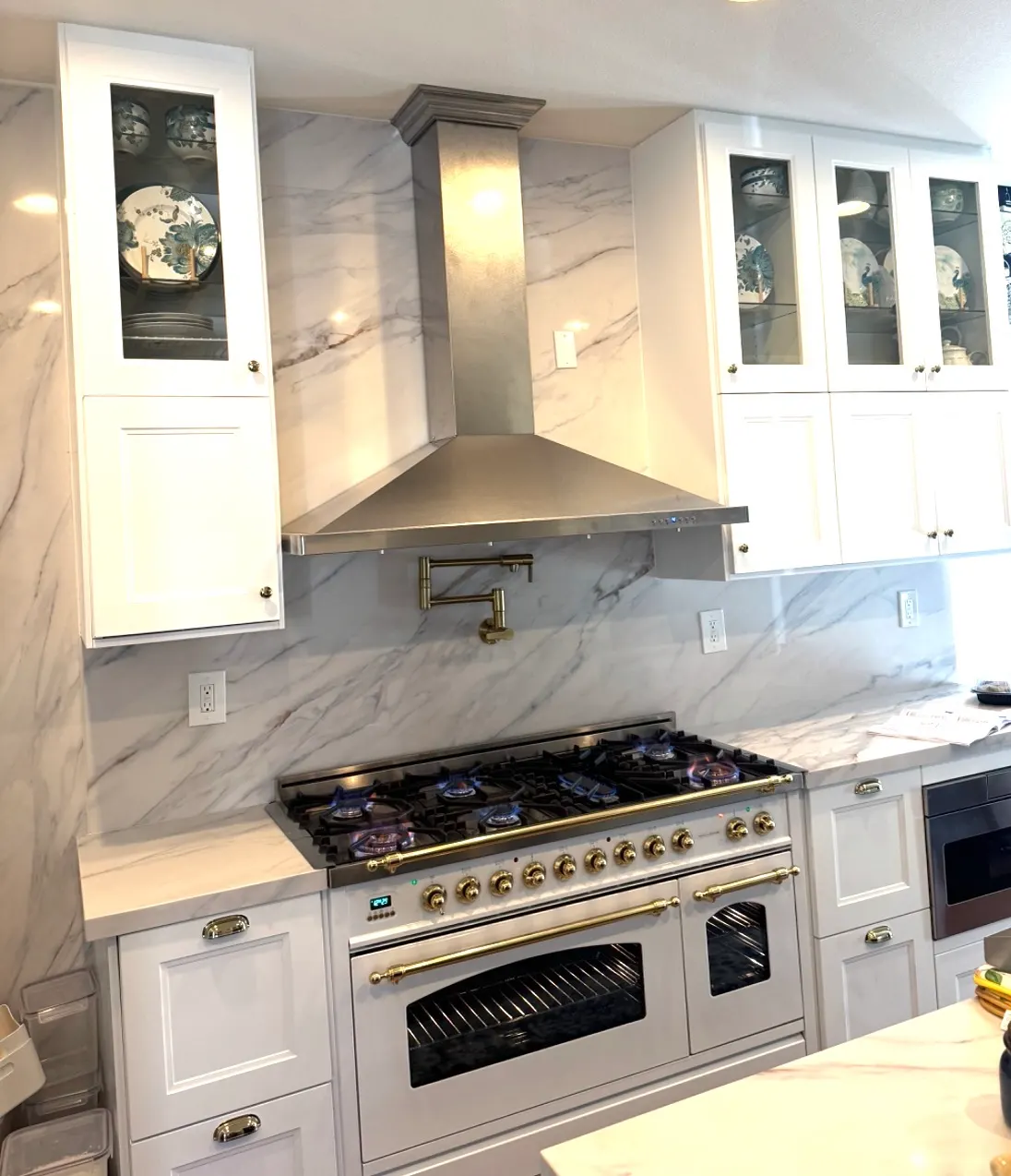 Luxurious kitchen features white marble backsplash and countertops, brushed brass pot filler, and a large professional range.