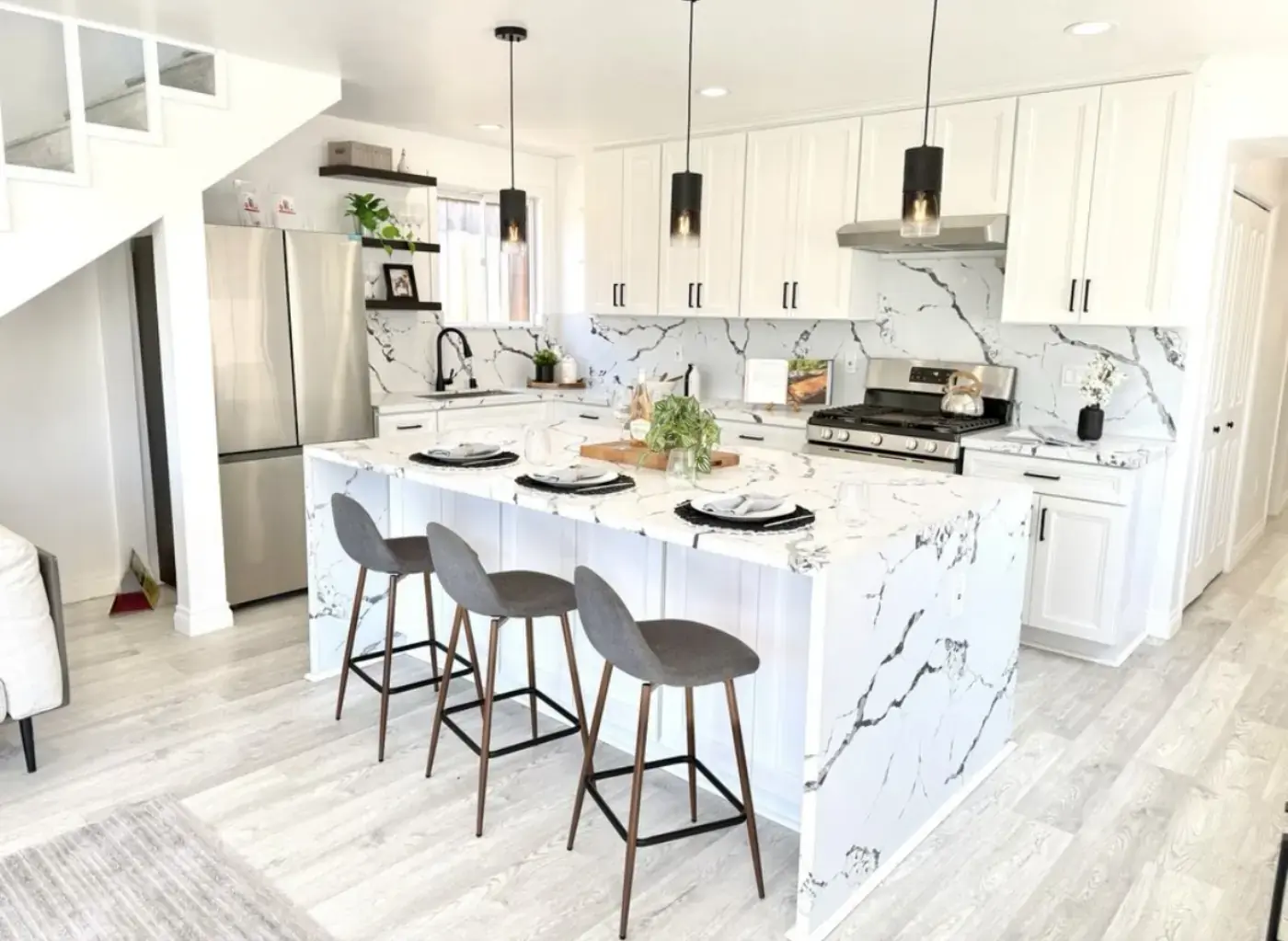 Open-concept white kitchen with dramatic marble waterfall island, modern pendant lights, and gray bar stools