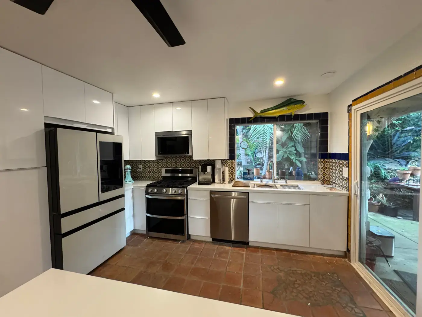 Compact modern kitchen with high-gloss white cabinets, decorative Mediterranean tile backsplash, and terracotta tile floor opening to a tropical patio